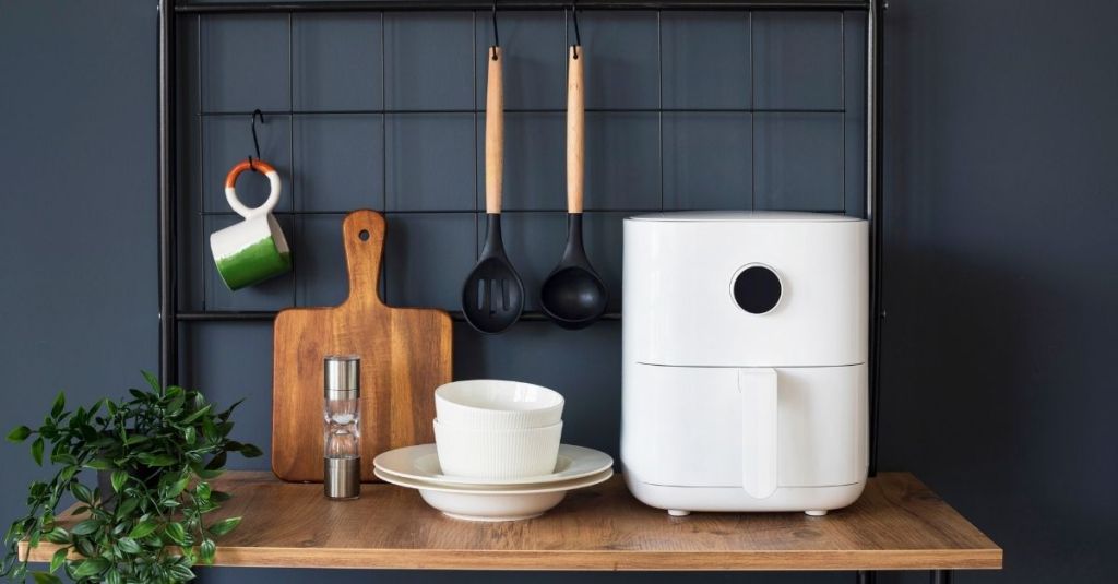 White air fryer on a wooden shelf with kitchen utensils and bowls, ideal for preparing crispy food with little oil.