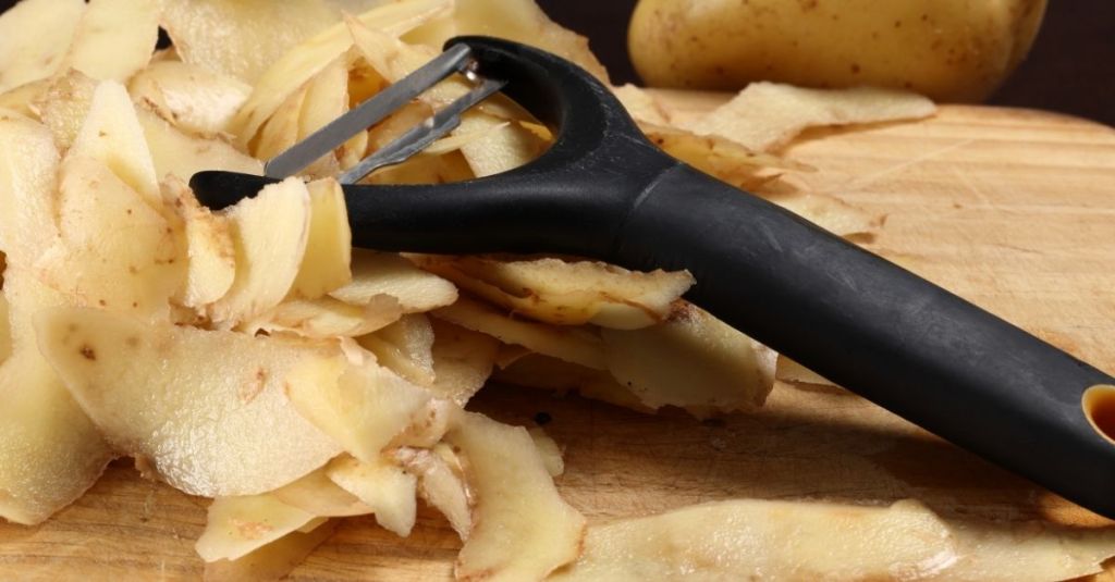Black-handled vegetable peeler with peeled potato skins on a wooden cutting board.