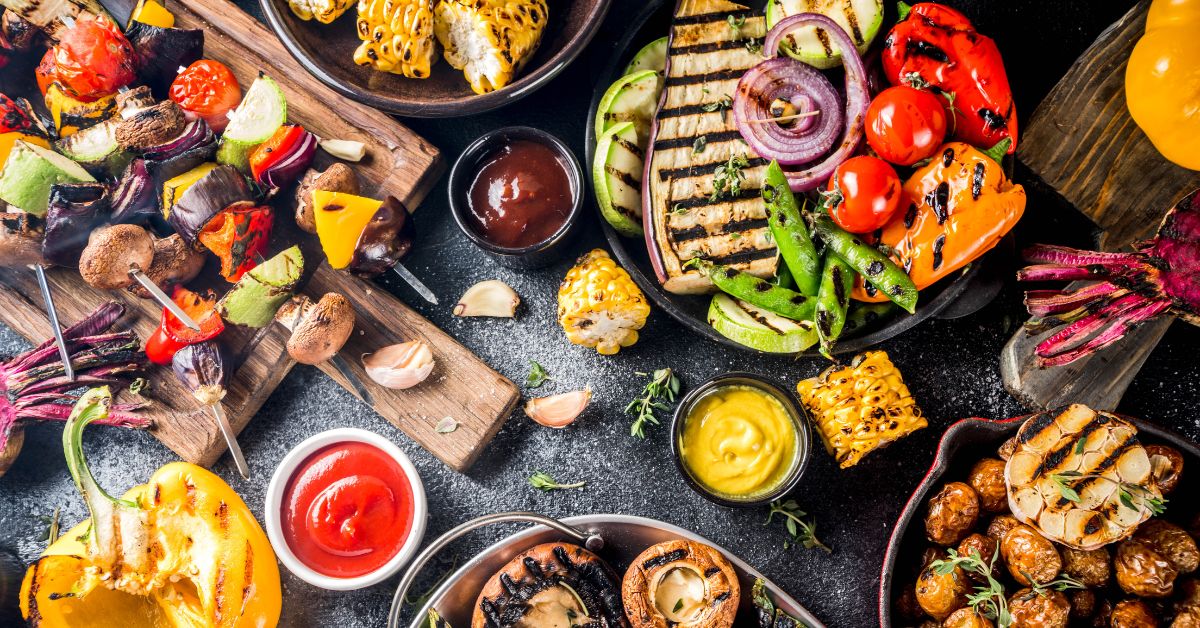 A top-down view of grilled plant-based dishes featuring colorful vegetables, skewers, sauces, and roasted garlic, ideal for vegan barbecues or healthy meal ideas.