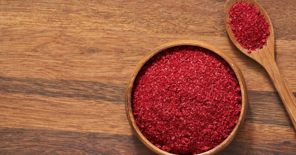 Ground sumac in a wooden bowl with a wooden spoon beside it, showcasing its vibrant red hue.
