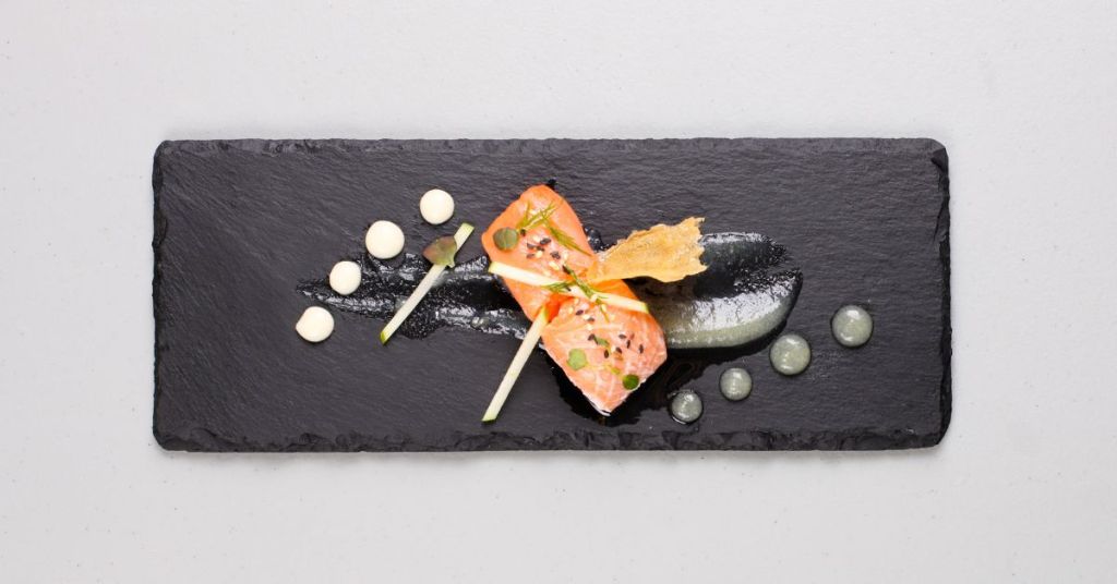 A gourmet dish featuring salmon, sauce drops, and garnishes, plated artistically on a black slate.