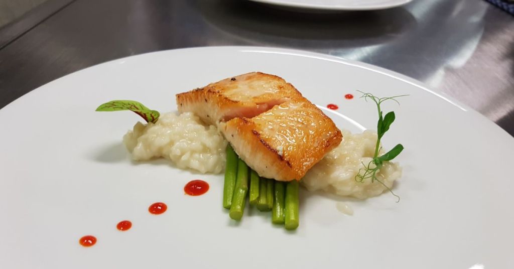 A gourmet dish featuring seared salmon, creamy risotto, green vegetables, and red gel accents, plated elegantly.