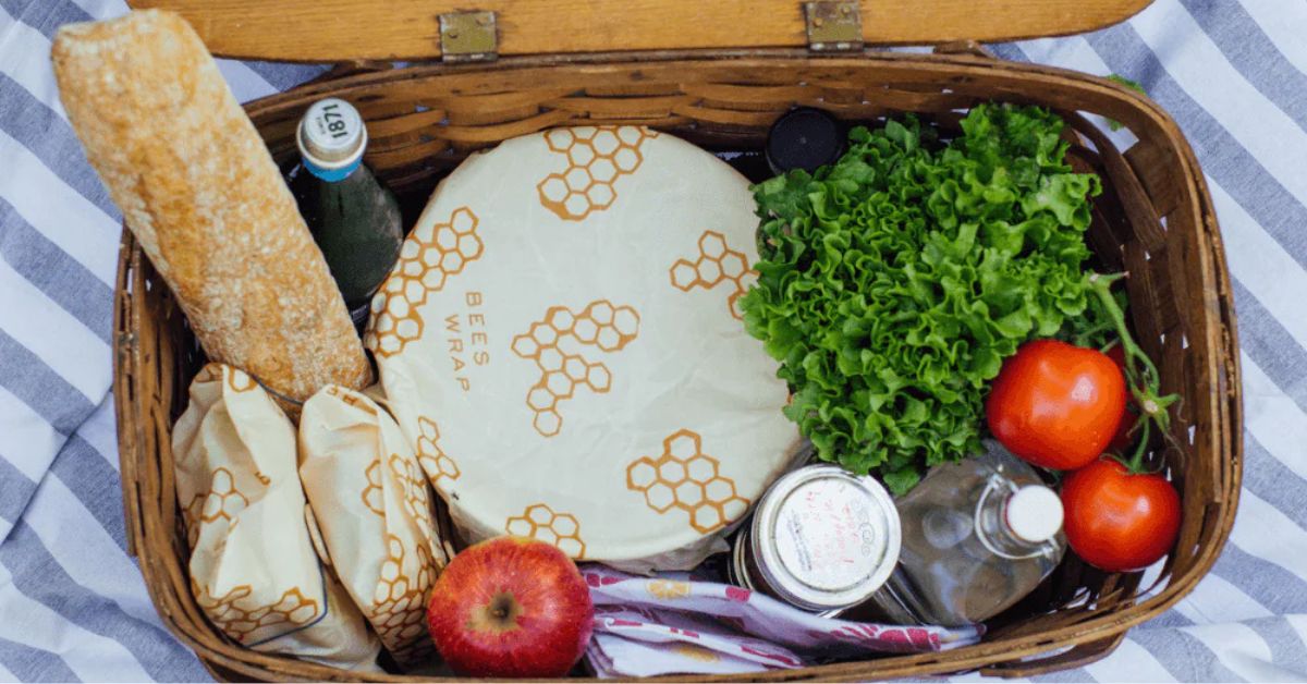 Eco-friendly reusable food wraps for sustainable storage. Wooden picnic basket filled with fresh produce, bread, and jars, using Bee's Wrap reusable wraps for sustainability.