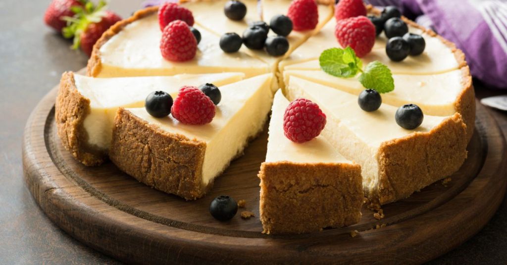 classic cheesecake with berry garnish A whole classic cheesecake with a golden crust, topped with fresh raspberries, blueberries, and mint leaves, displayed on a rustic wooden board with slices ready to serve.