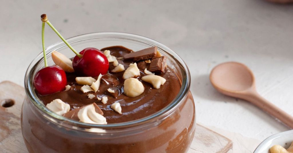 Chocolate avocado mousse in a glass jar, topped with cherries, macadamia nuts, and chocolate pieces. Dairy-free chocolate mousse made with avocado, garnished with cherries and chopped nuts.