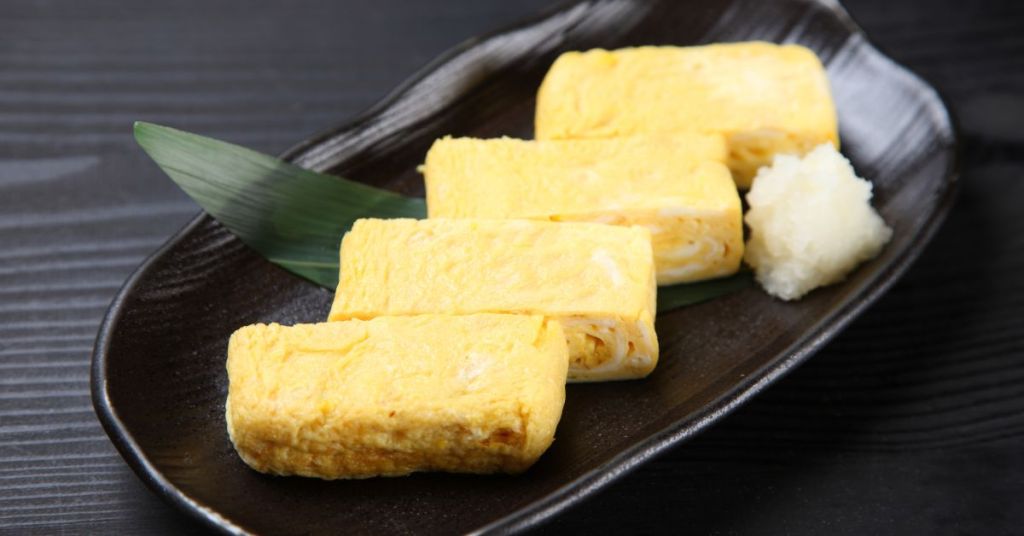 Japanese tamagoyaki, a delicate omelet in a rolled egg preparation made with precision, served with grated daikon on a black plate.