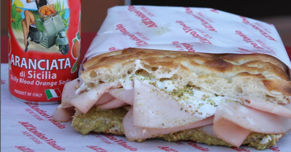 Close-up of the La Paradiso sandwich from All’Antico Vinaio with layers of mortadella, stracciatella, and pistachio cream, accompanied by a can of Sicilian blood orange soda.