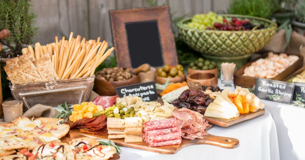 A rustic outdoor charcuterie table with a variety of cheeses, cured meats, pickles, breadsticks, and fresh fruits, styled for a large gathering.