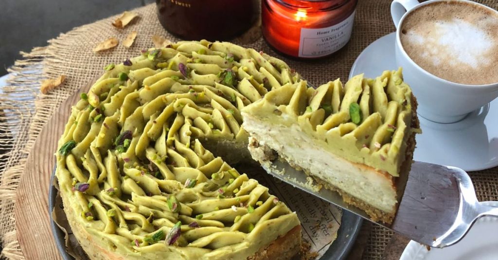 pistachio cheesecake with intricate piping Whole pistachio cheesecake topped with elegant piped pistachio cream and crushed pistachios, with a slice being served alongside coffee.