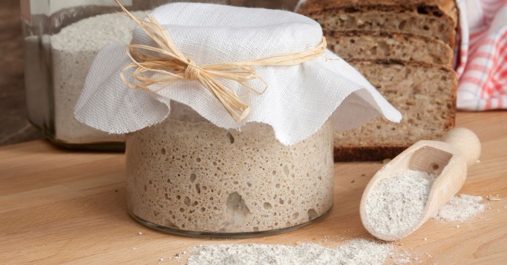 A sourdough starter bubbling in a jar, covered with a cloth and raffia ribbon, surrounded by rustic bread and flour.A sourdough starter bubbling in a jar, covered with a cloth and raffia ribbon, surrounded by rustic bread and flour.