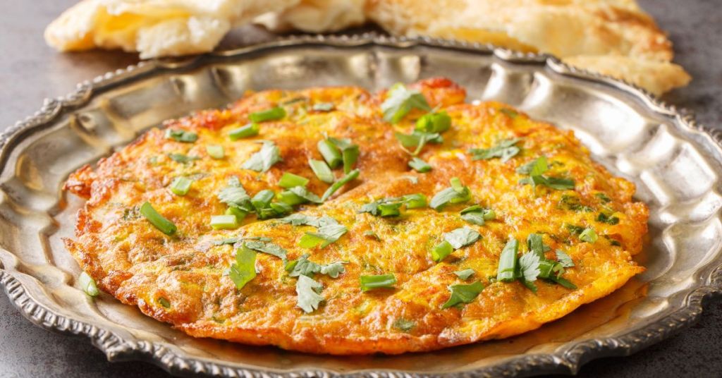 A golden Indian omelet infused with spices and garnished with cilantro, served on a traditional silver plate.