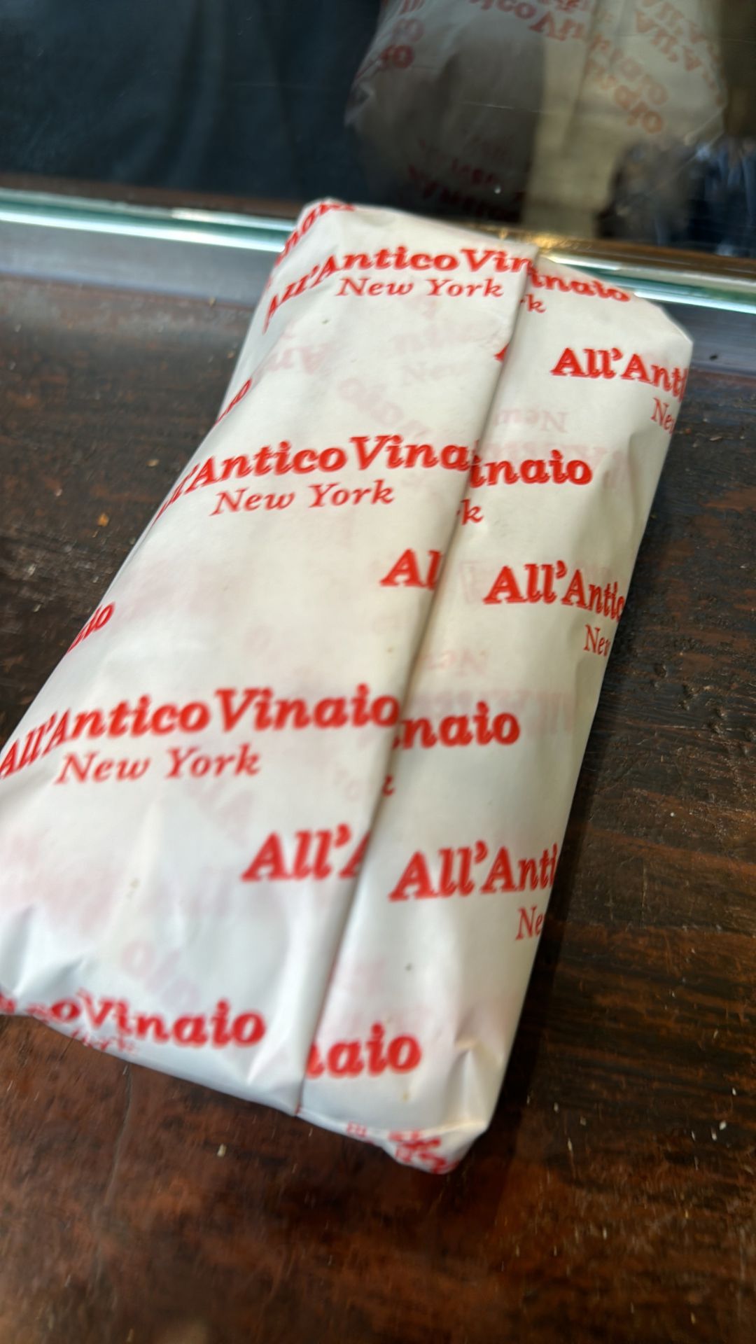 Close-up of a sandwich wrapped in All’Antico Vinaio’s signature white paper with red branding, resting on a wooden countertop.