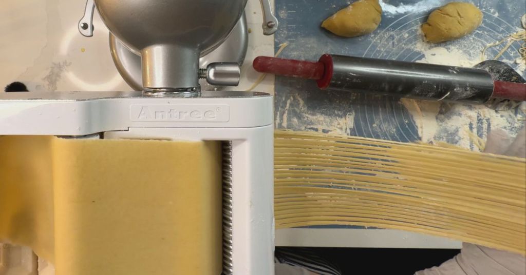 Fresh pasta dough being rolled and cut into linguini strands with a KitchenAid pasta machine attachment.