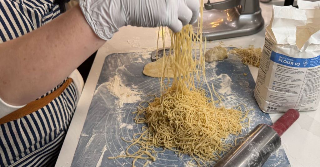 Hands lifting freshly made pasta strands on a floured surface, with a KitchenAid mixer and flour in the background.